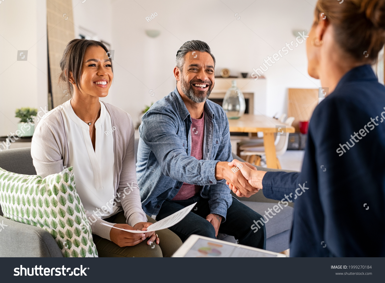 Mature indian man shaking hands with financial advisor at home. Happy smiling couple greeting broker with handshake at home. Multiethnic mid adult man and hispanic woman sealing a contract. #1999270184