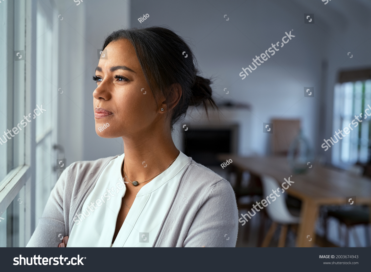 Mature african woman looking outside window with uncertainty. Thoughtful mid adult woman looking away through the window while thinking about her future business after pandemic. Doubtful lady at home. #2003674943