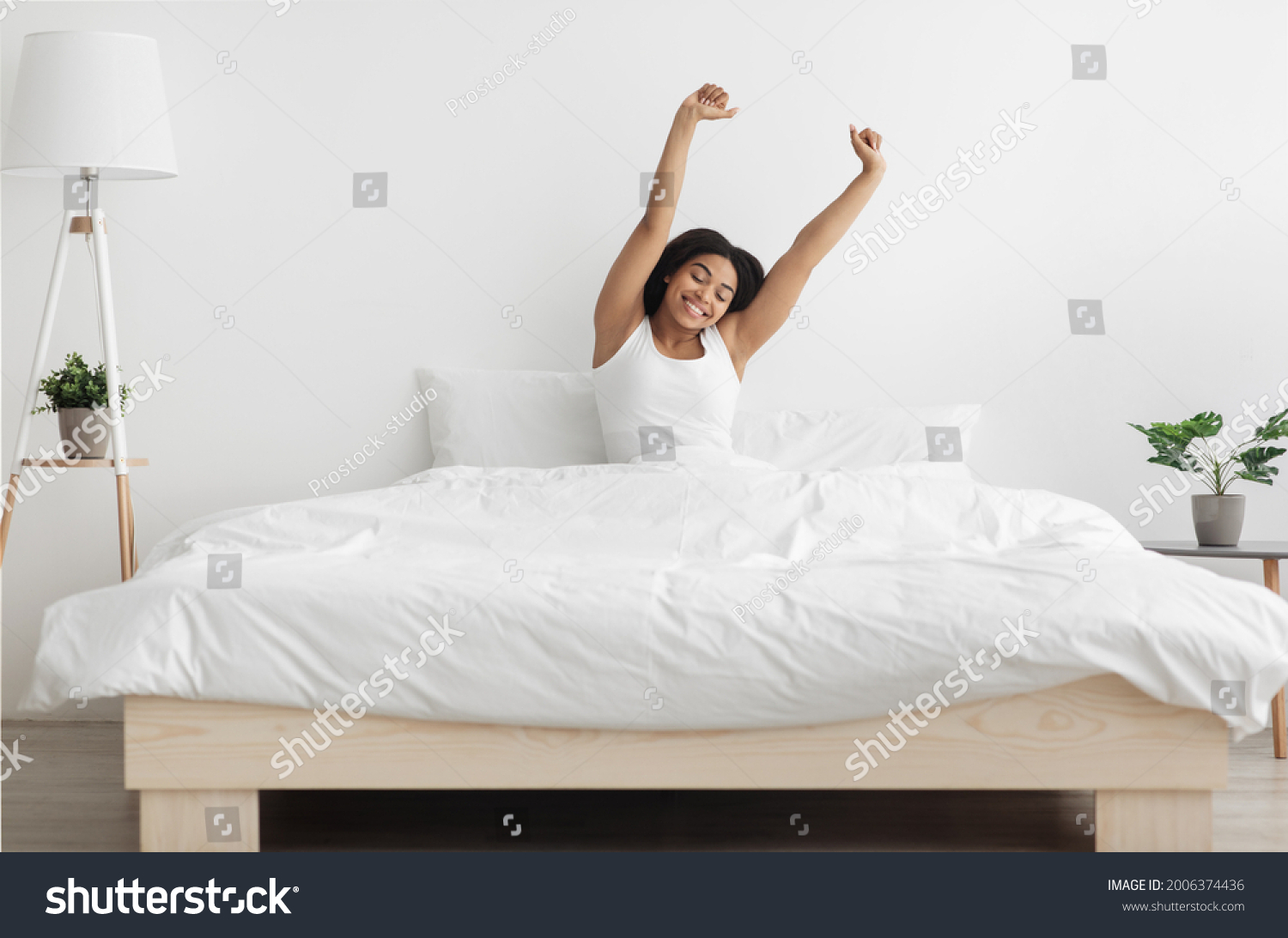 Good start of new day. Well-slept african american lady stretching arms after waking up, feeling happy and full of vitality. Young woman sitting in comfortable bed in coy bedroom interior #2006374436
