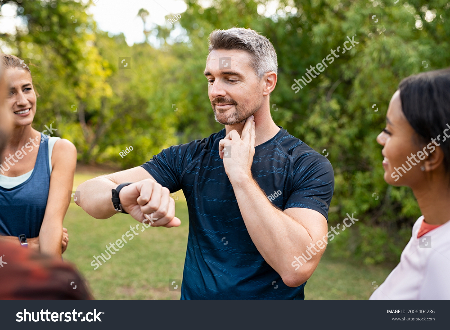 Mature tired man checking pulse after workout. Handsome middle aged man standing with group of friends measuring heart rate pulse on his neck and looking sport watch. Guy times the pulsations at park. #2006404286