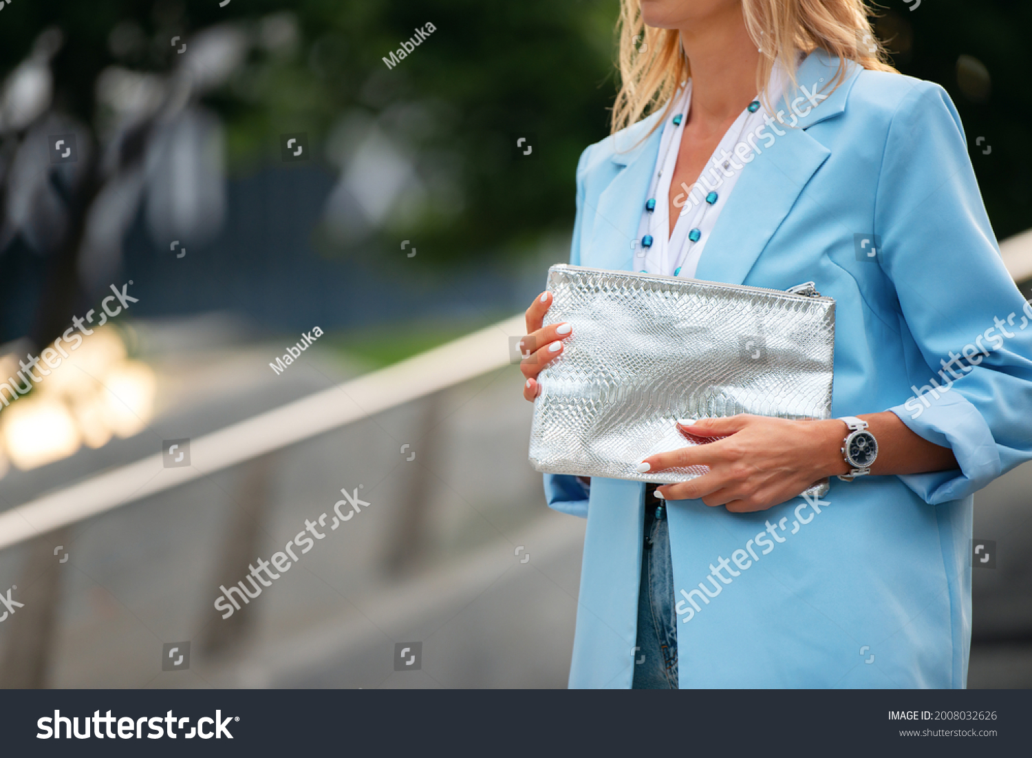 Women's silver clutch bag with snakeskin texture. Fragment of the body of a model in a summer blue jacket, white blouse. Business attire and beads. #2008032626