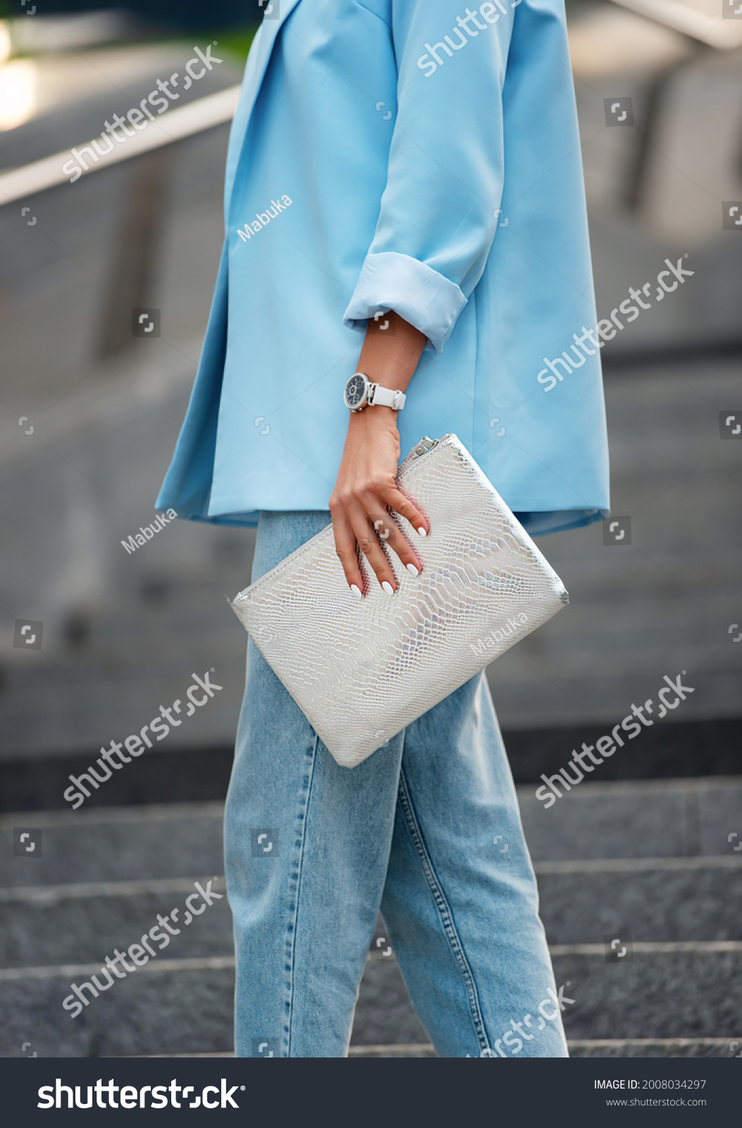 Women's silver clutch bag with snakeskin texture. A fragment of the body of a model in a summer blue blazer with rolled-up sleeves and blue jeans. Business style clothing. #2008034297