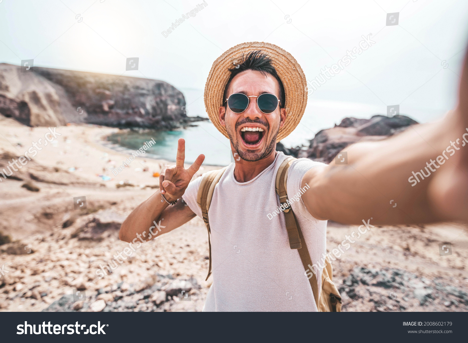 Young man with backpack taking selfie portrait on a mountain - Smiling happy guy enjoying summer holidays at the beach - Millennial showing victory hands symbol to the camera - Youth and journey  #2008602179