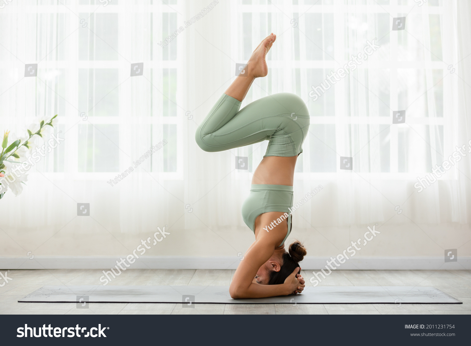 Side view of Asian woman wearing sportwear doing Yoga exercise in front of windows,Yoga Forearm Stand pose or Pincha Mayurasana.Calm healthy young female breathing and meditate yoga self care at home #2011231754