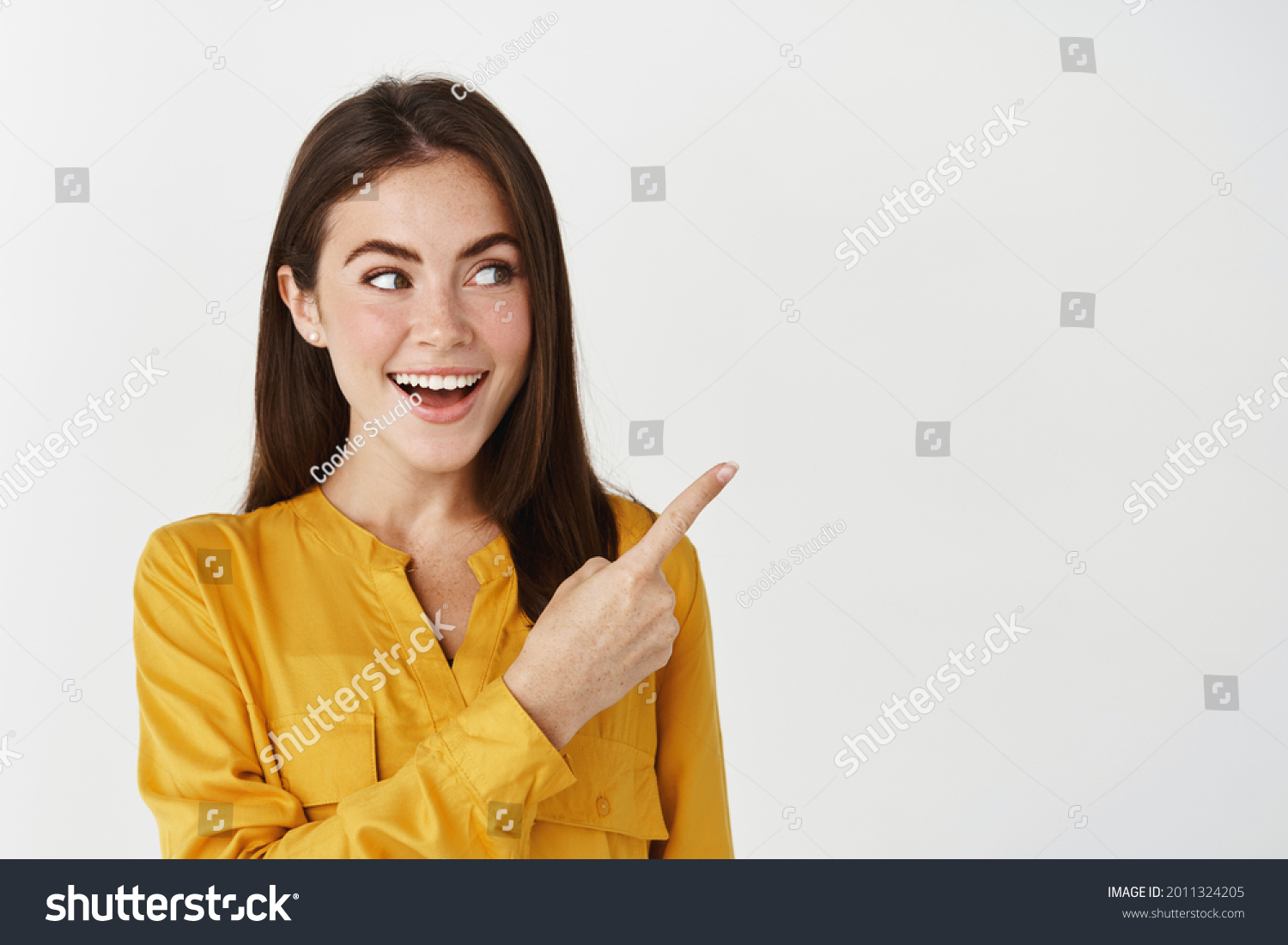Close-up of lady smiling, pointing and looking right with surprised face, standing on white background #2011324205