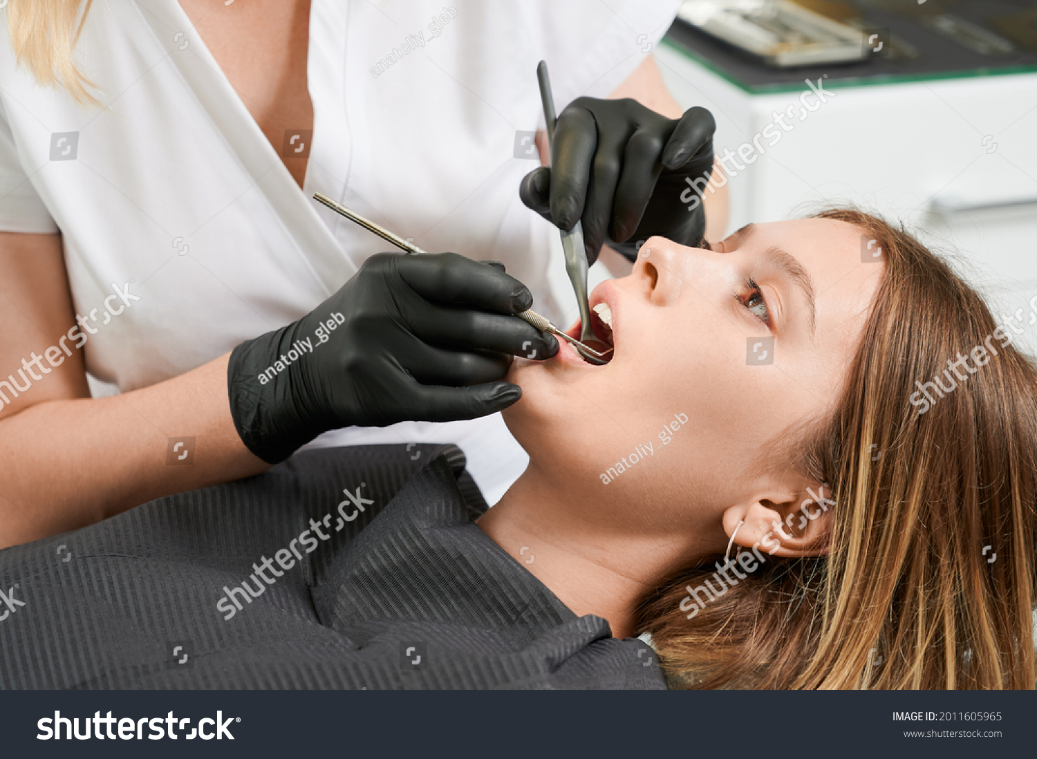 Female dentist in black sterile gloves examining patient teeth with dental explorer and mirror. Young woman lying in dental chair while having prophylactic examination. Concept of dentistry. #2011605965