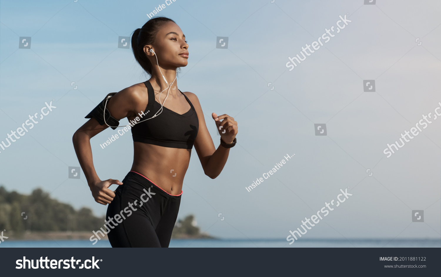 Side view shot of young woman in sportswear jogging on beach. African-american female jogger runner running outdoors. Active lifestyle concept #2011881122