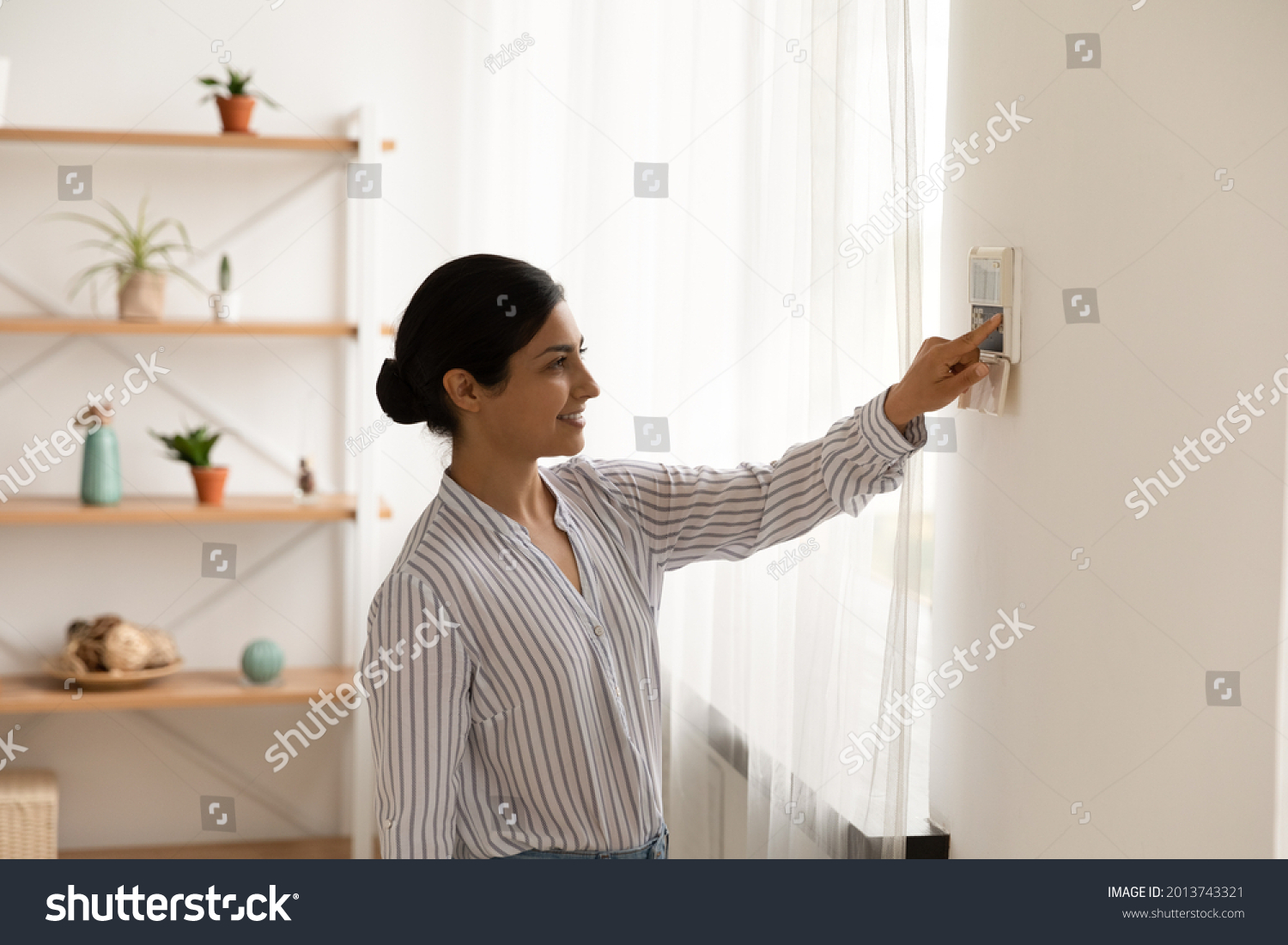 Smiling Indian woman using modern smart home system, controller on wall, positive attractive young female switching temperature on thermostat or activating security alarm in apartment #2013743321