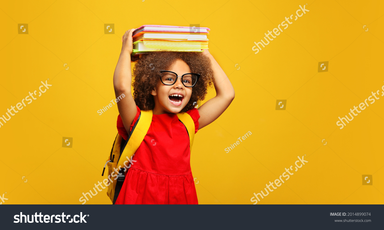 funny smiling Black child school girl with glasses hold books on her head. Yellow background #2014899074