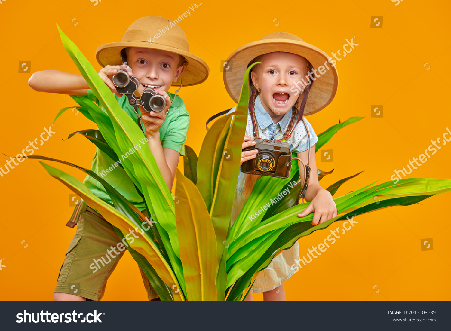 Young travelers, a girl and a boy, explore nature, they look out from behind palm leaves with binoculars and a camera. Summer vacation, tourism. Studio portrait on a yellow background.  #2015108639