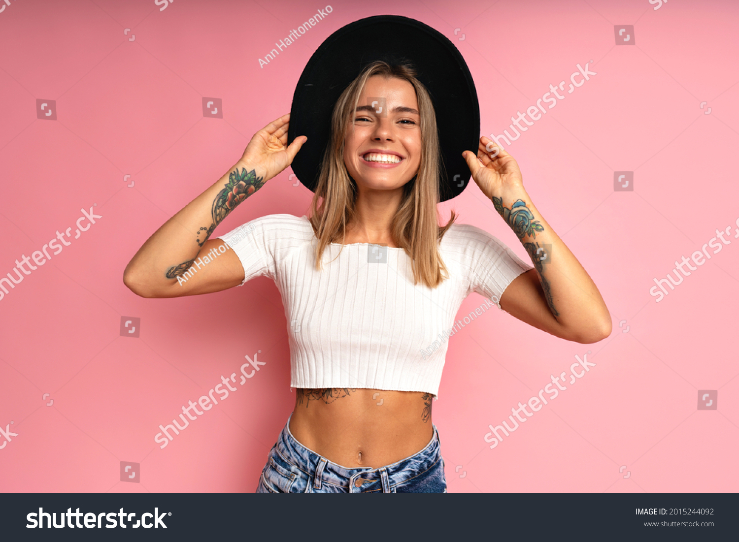 Cheerful stylish woman posing at studio pink background, wearing crop top blue jeans, black fedora. Blonde hairs and tattos. #2015244092