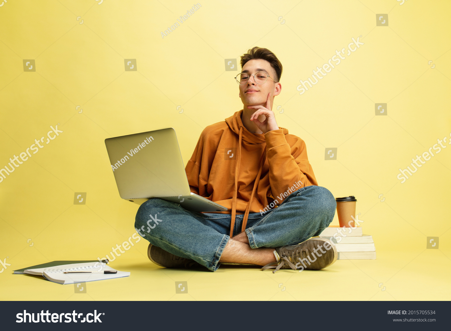 Studying, doing homework. One young smiling caucasian man, student in glasses sits on floor with laptop isolated on yellow studio background. Education, studying and student life concept. #2015705534