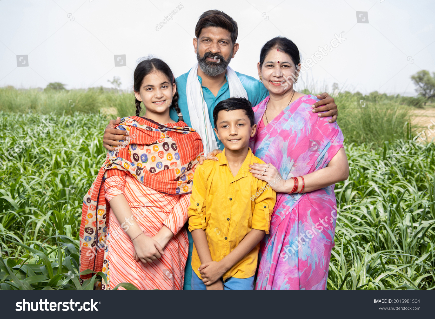 Portrait of happy rural indian family standing in agriculture field. Parents with their children, farmer husband wife with daughter and son. Beard man wearing kurta and woman wearing sari,  #2015981504