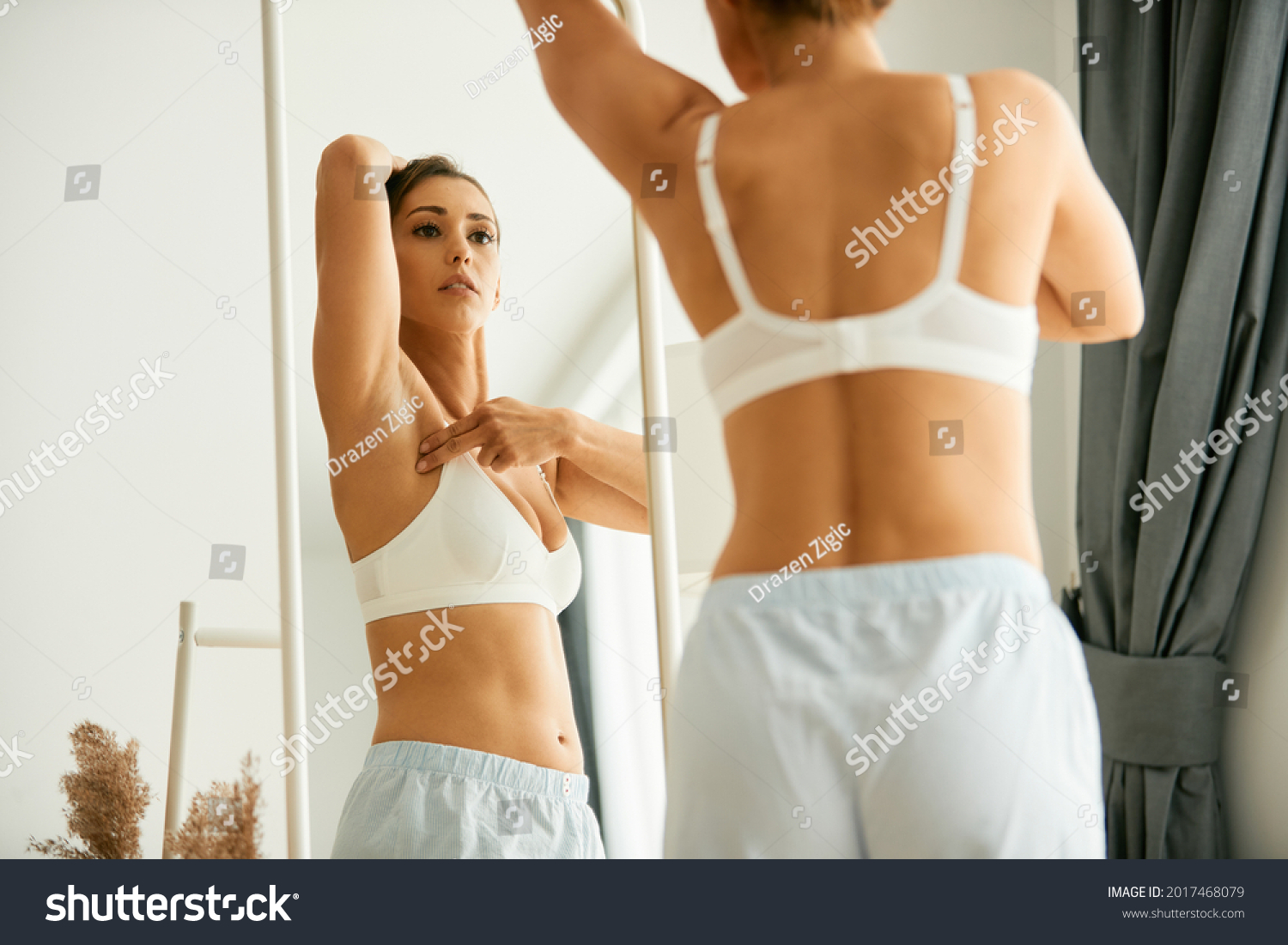 Cancer aware young woman looking herself in a mirror while doing breast self-examination at home.  #2017468079
