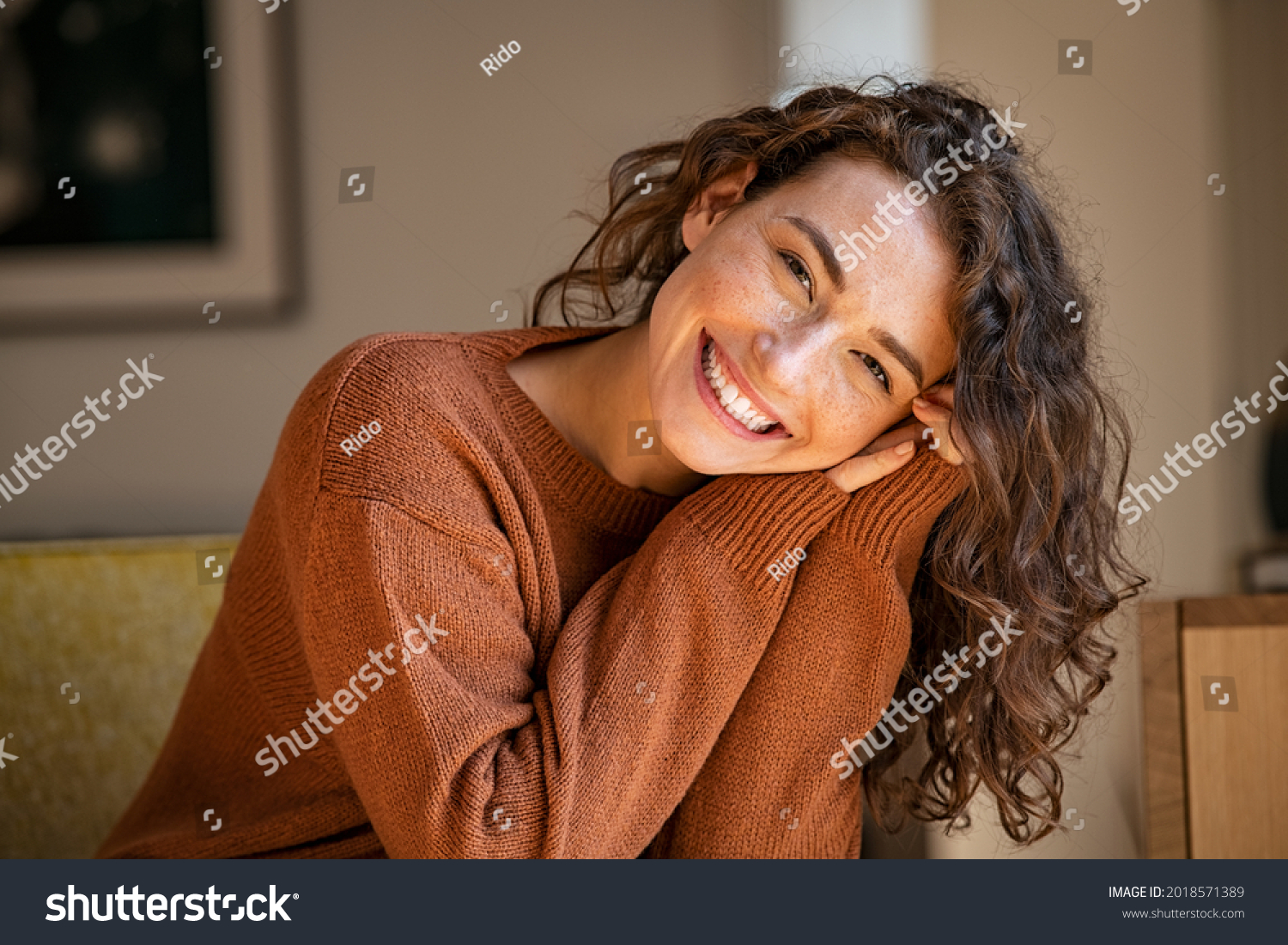 Happy young woman sitting on sofa at home and looking at camera. Portrait of comfortable woman in winter clothes relaxing on armchair. Portrait of beautiful girl smiling and relaxing during autumn. #2018571389