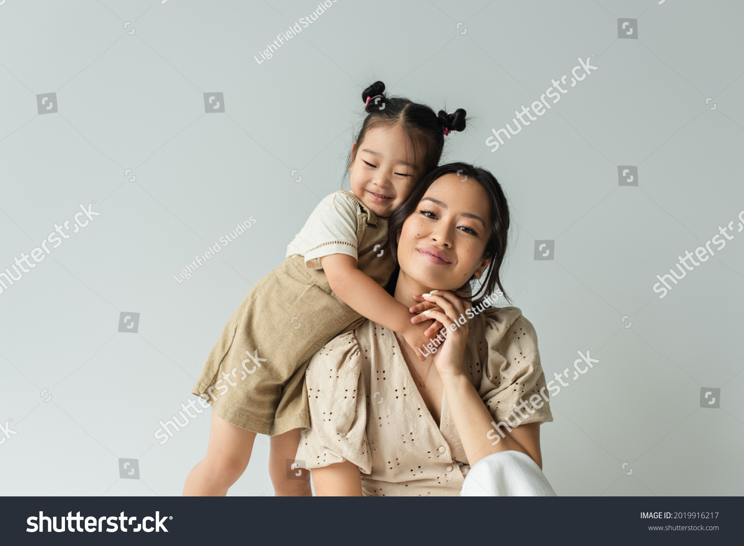 cheerful asian toddler daughter hugging happy mother isolated on gray #2019916217