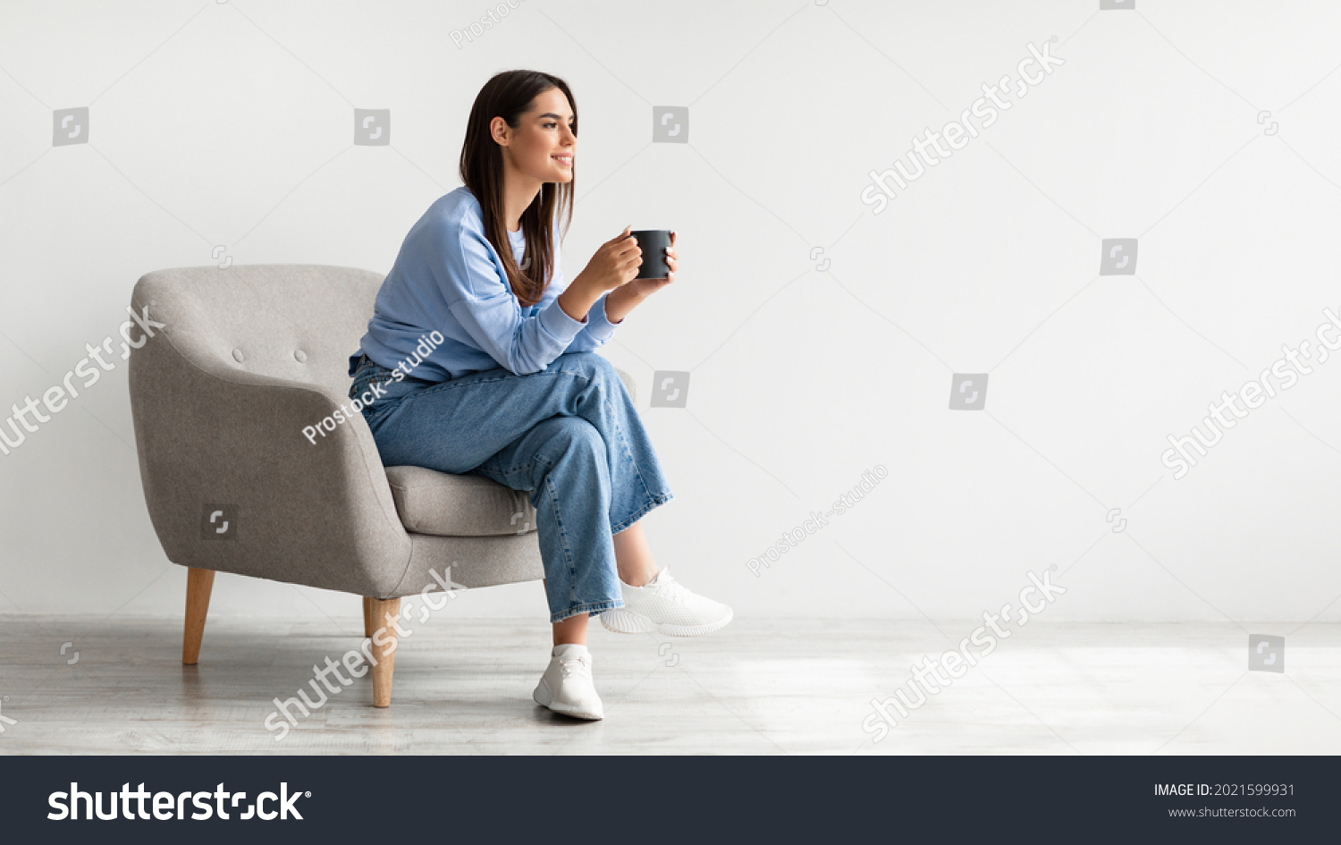 Full length of young woman drinking hot coffee in cozy armchair against white studio wall, banner design with free space. Peaceful lady having relaxing day, chilling on lazy morning #2021599931