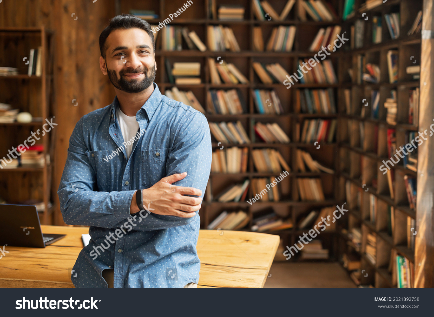 Smiling bearded indian businessman stands near desk and looks at the camera. Young positive male student in library with bookshelves on background. Proud and successful mixed-race small business owner #2021892758