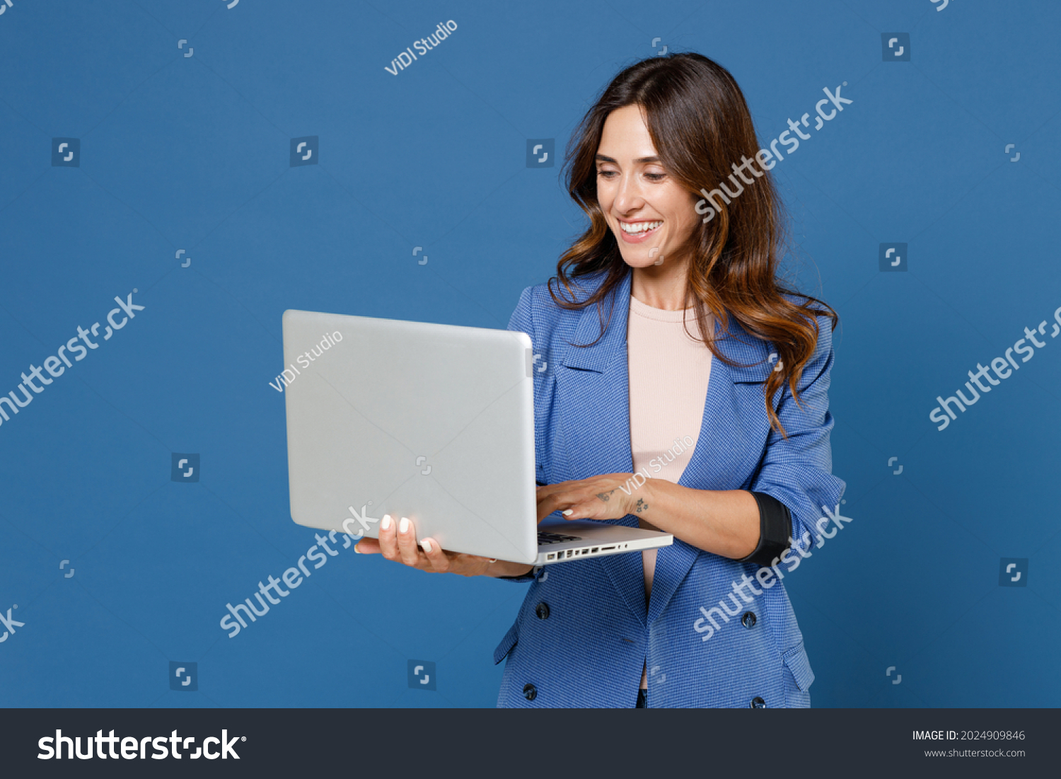 Smiling cheerful funny beautiful attractive young brunette woman 20s wearing basic casual jacket standing working on laptop pc computer isolated on bright blue colour background studio portrait #2024909846