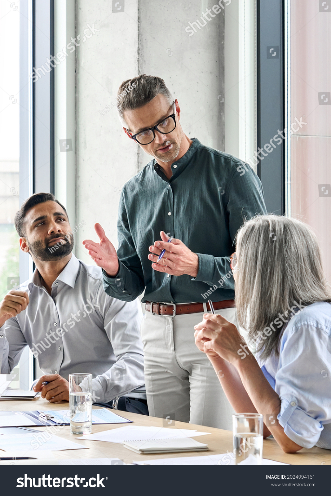 Mature businessman leader mentor talking to diverse colleagues team listening to caucasian ceo. Multicultural professionals project managers group negotiating in boardroom at meeting. Vertical. #2024994161