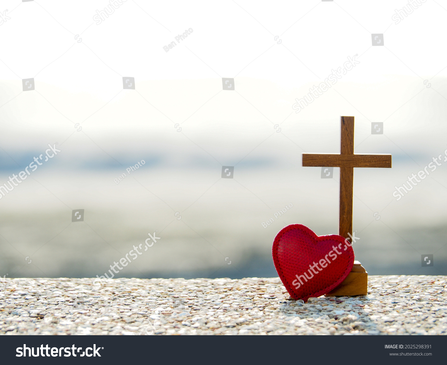 Red heart with wooden Christian cross on gravel floor in morning light, beach sea as background. Jesus love you. Faith hope believe in God. Believe in salvation. Christianity background concept. #2025298391