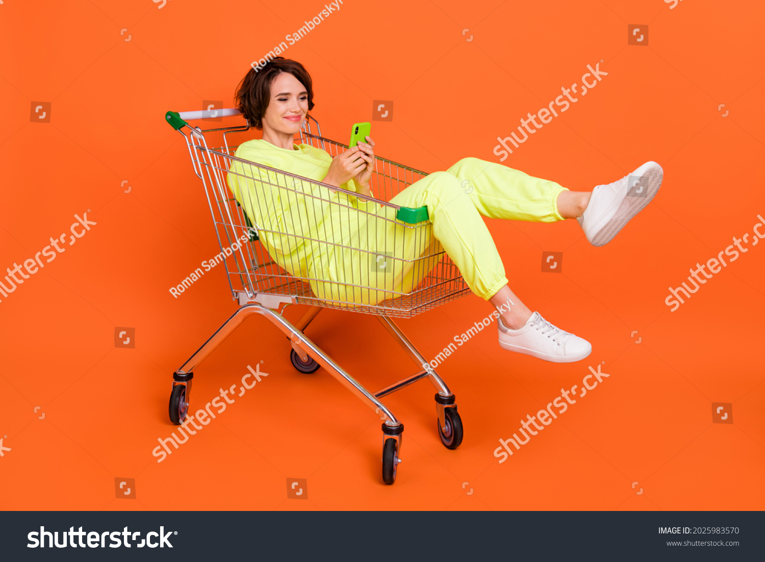 Portrait of attractive cheery funny focused girl sitting in shopping cart chatting blogging isolated over bright orange color background #2025983570