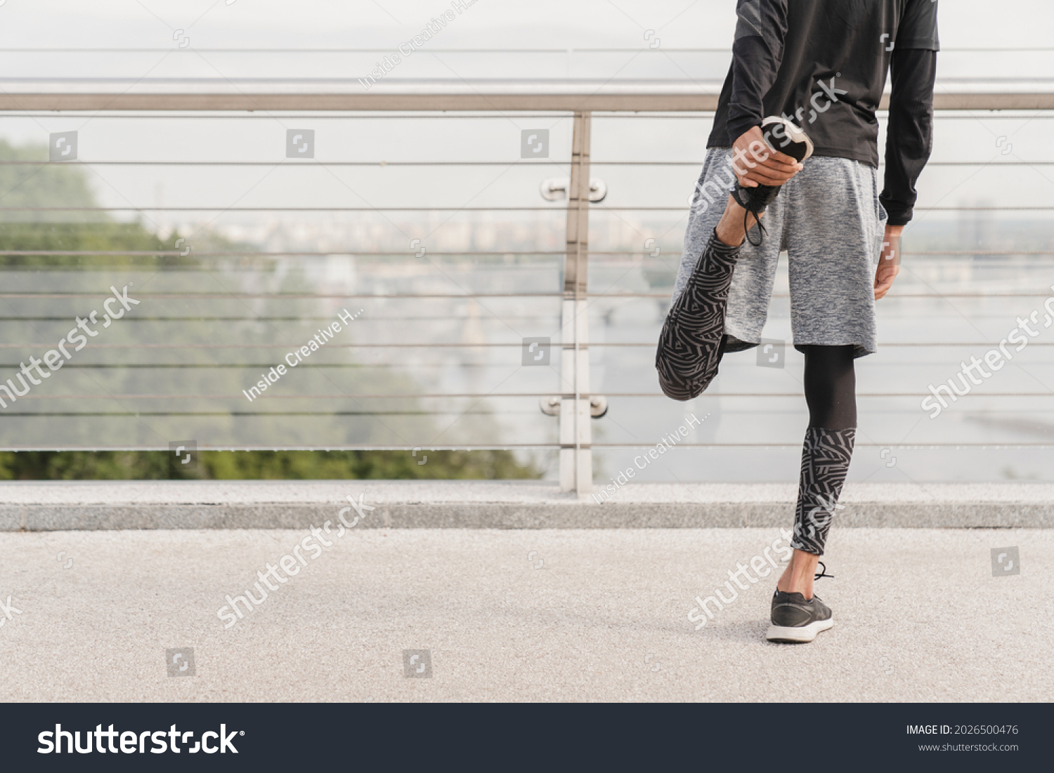 Young fit slim sportsman male athlete stretching his legs before jogging running on city bridge in urban area in sporty fitness clothes and trainers. Warming-up exercises outdoors. #2026500476