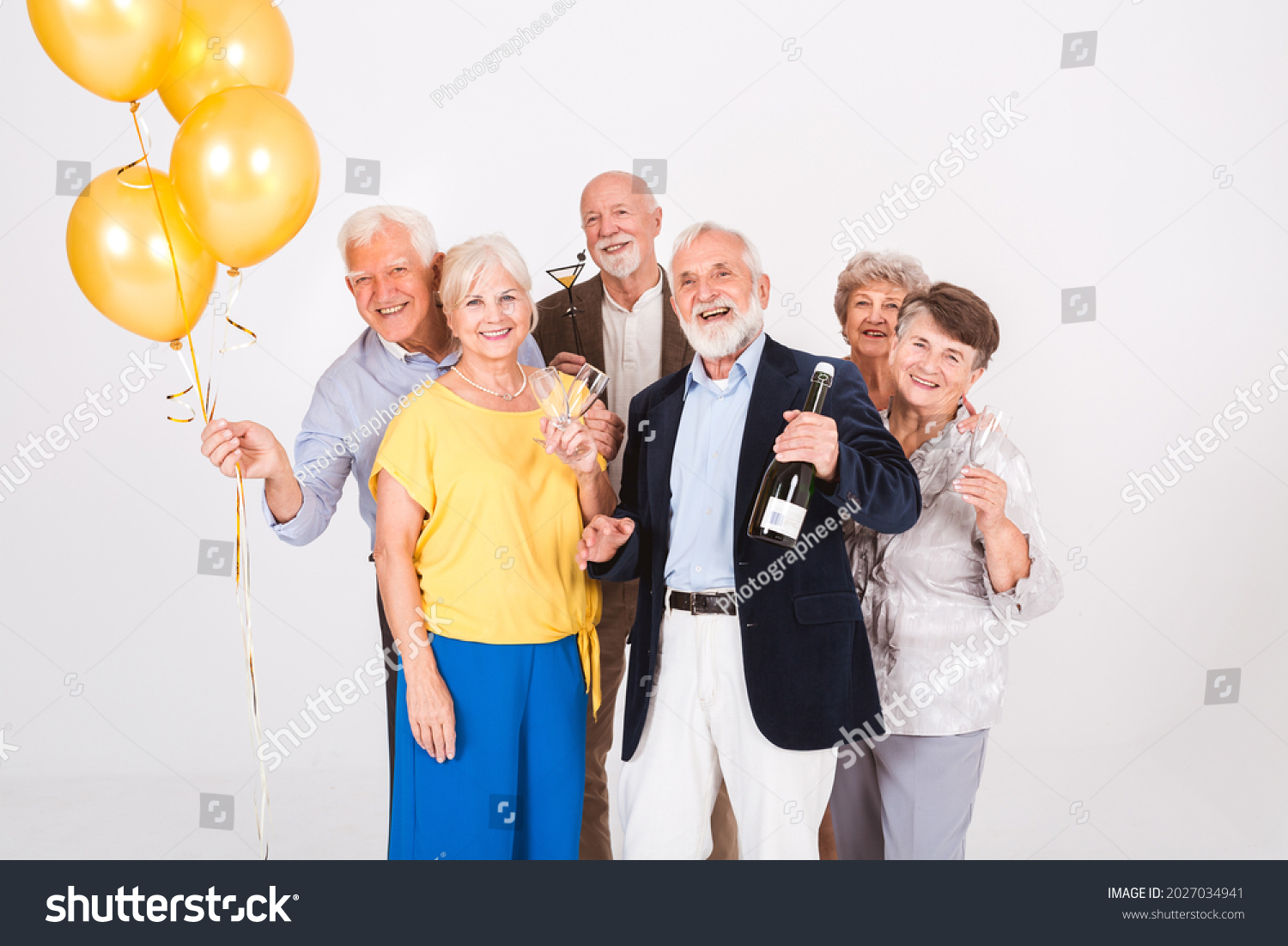 Group of happy senior friends holding yellow balloons and standing in white interior #2027034941