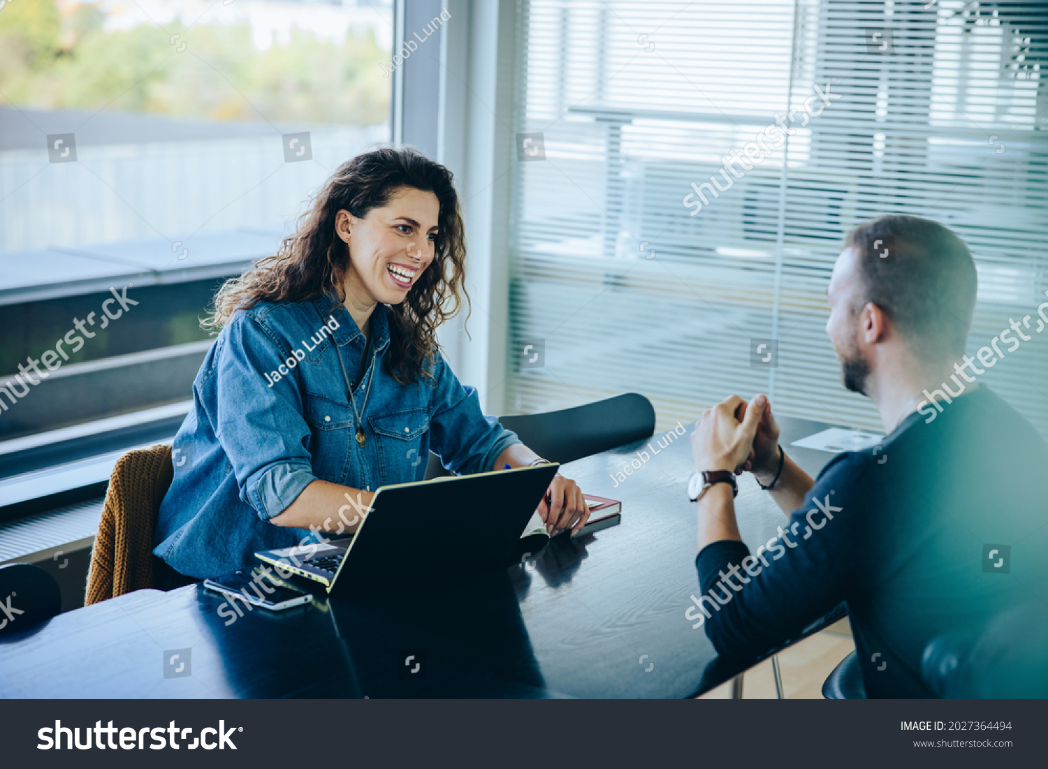 Smiling businesswoman taking interview of a job applicant. Friendly recruitment manager interviewing young man in office boardroom. #2027364494