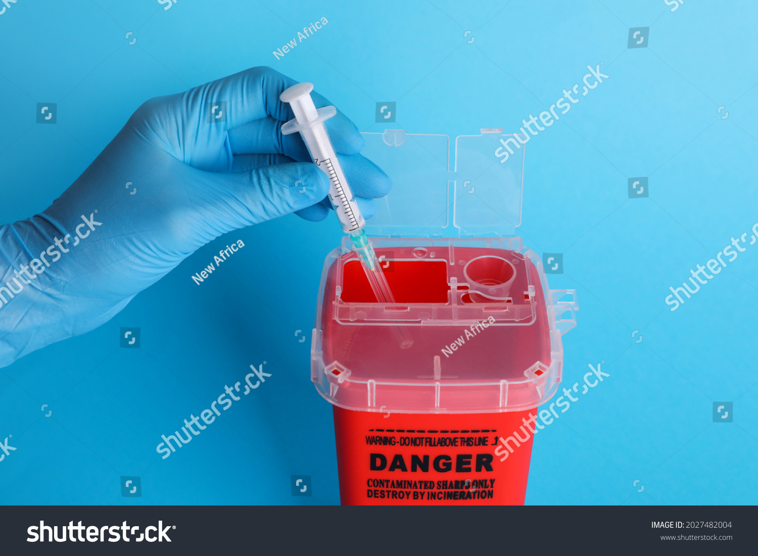 Doctor throwing used syringe into sharps container on light blue background, closeup #2027482004