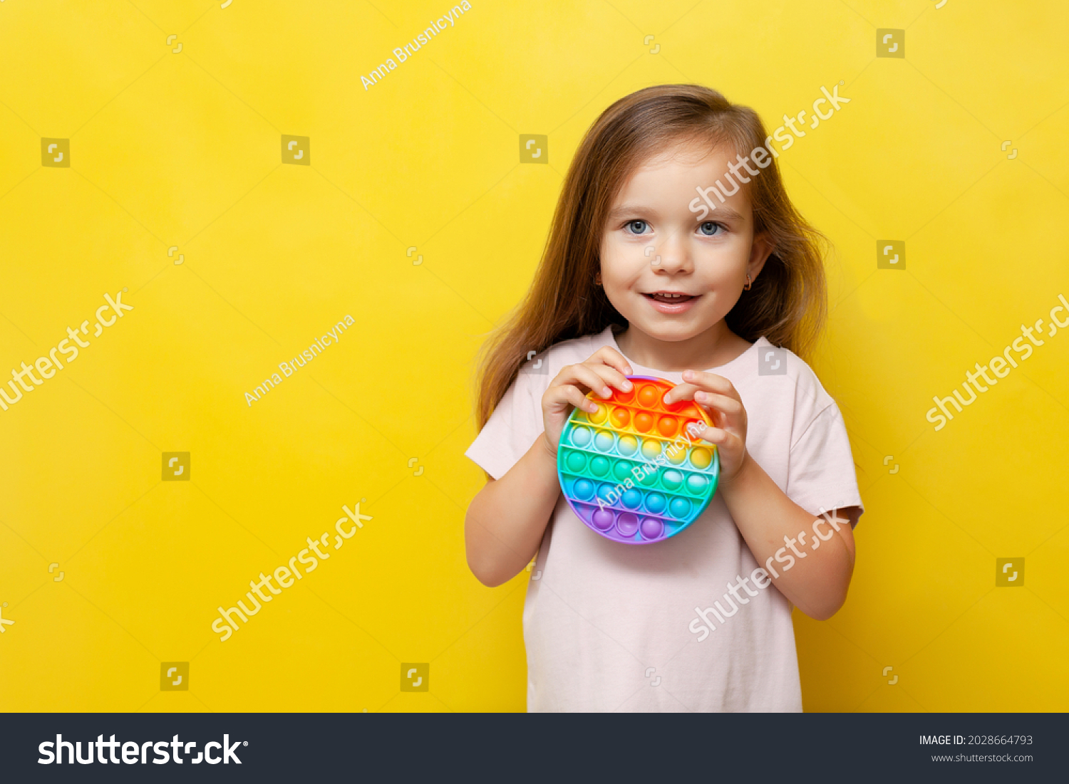 A beautiful cute girl holds a colored pop it toy in her hands and smiles. Yellow background. Anti-stress, emotions, good mood. A place for text. Trend #2028664793
