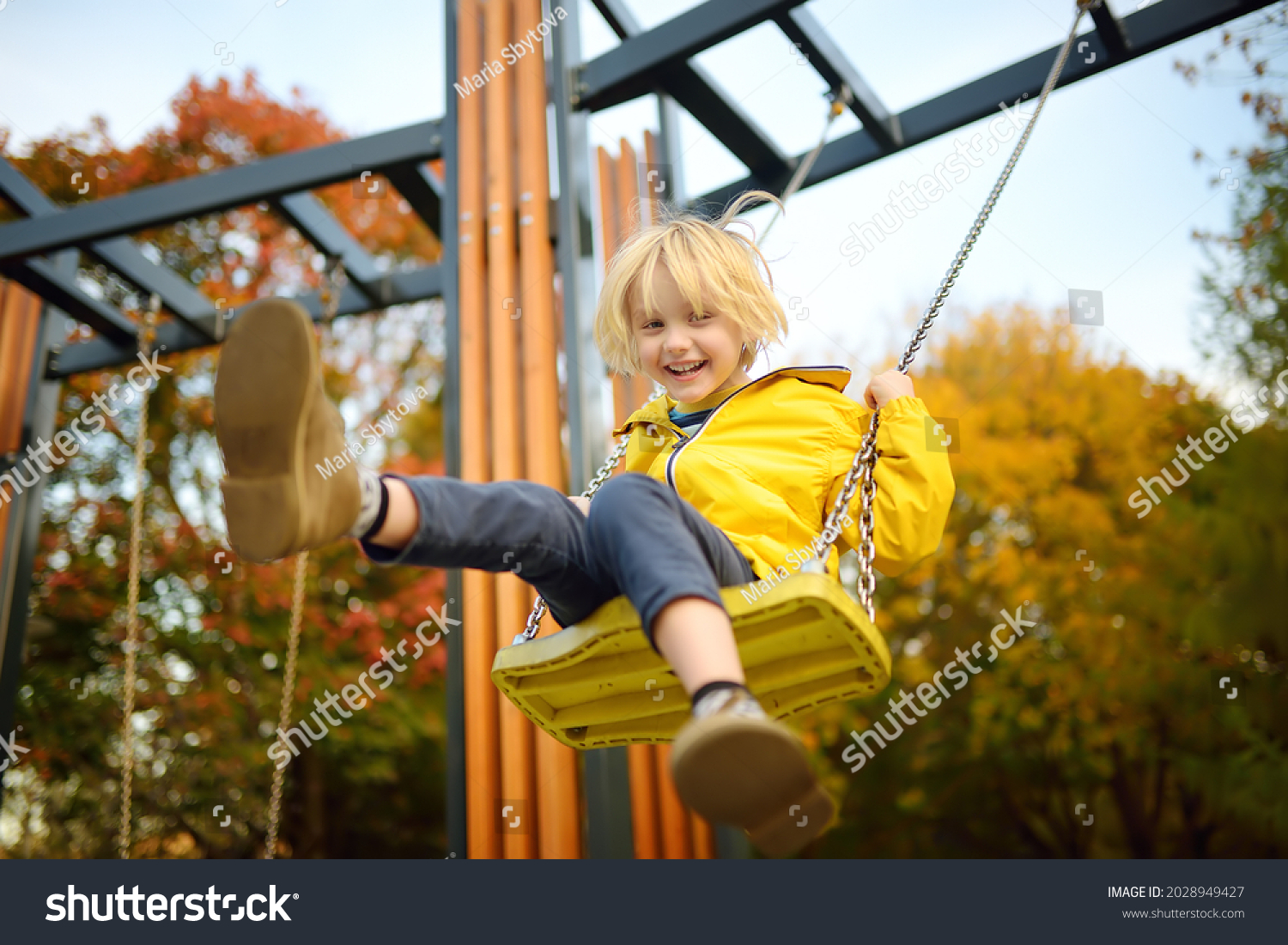Little boy having fun on a swing on the playground in public park on autumn day. Happy child enjoy swinging. Active outdoors leisure for child in city #2028949427