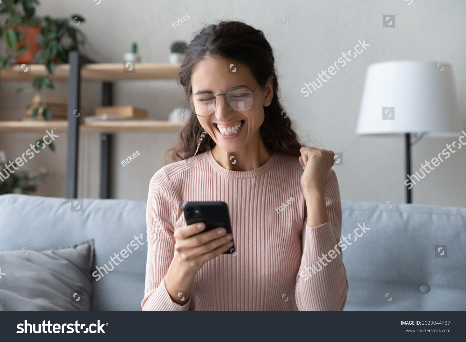 Attractive happy Latina woman in glasses sit on sofa at home holding smartphone read sms notification great news looking excited overjoyed cant believe in luck enjoy moment of victory. Success concept #2029044737