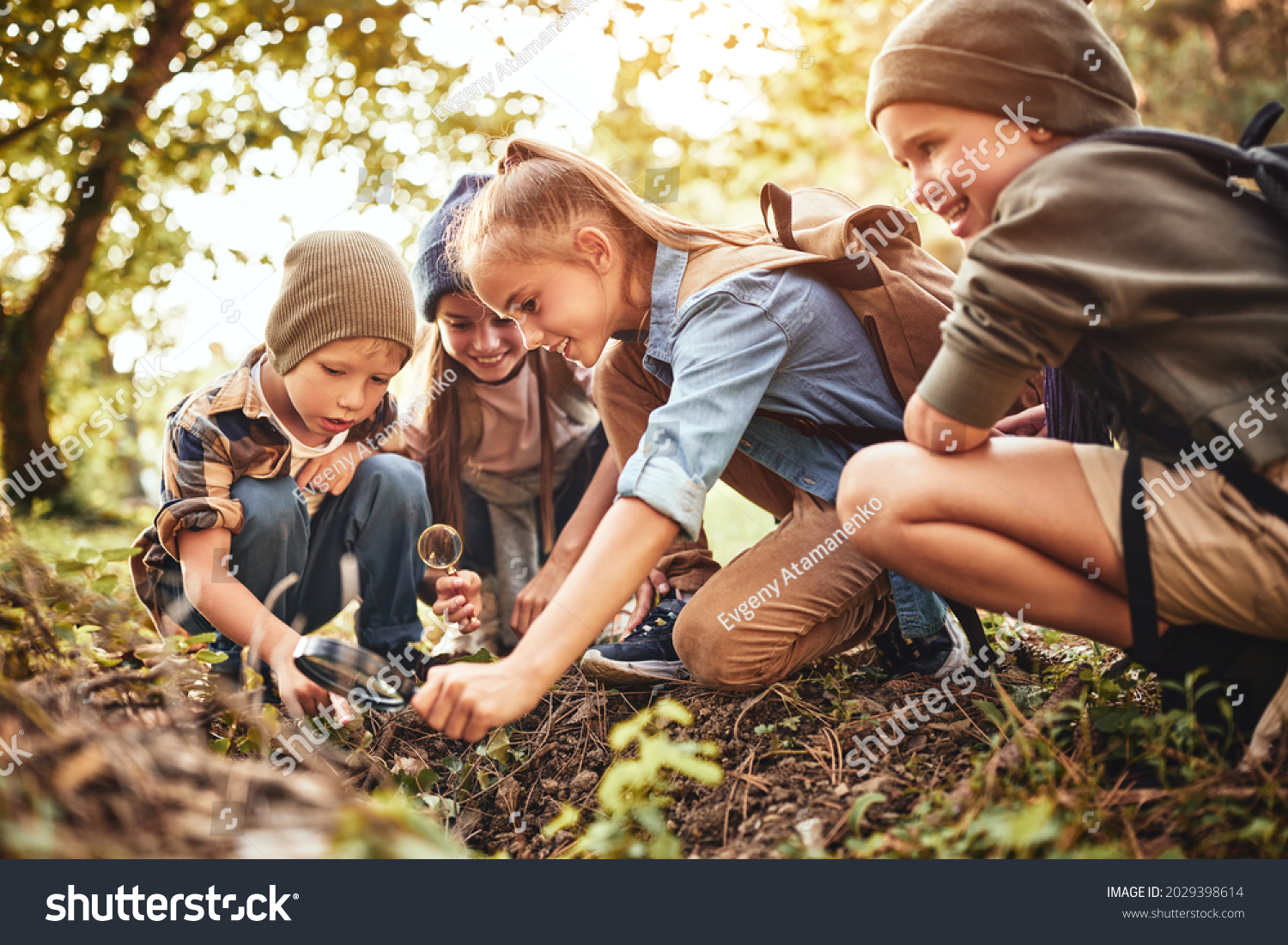 Happy children boys and girls in casual clothes with backpacks making bonfire with magnifying glass together in green forest during school camping activity on sunny day, smiling kids exploring nature #2029398614