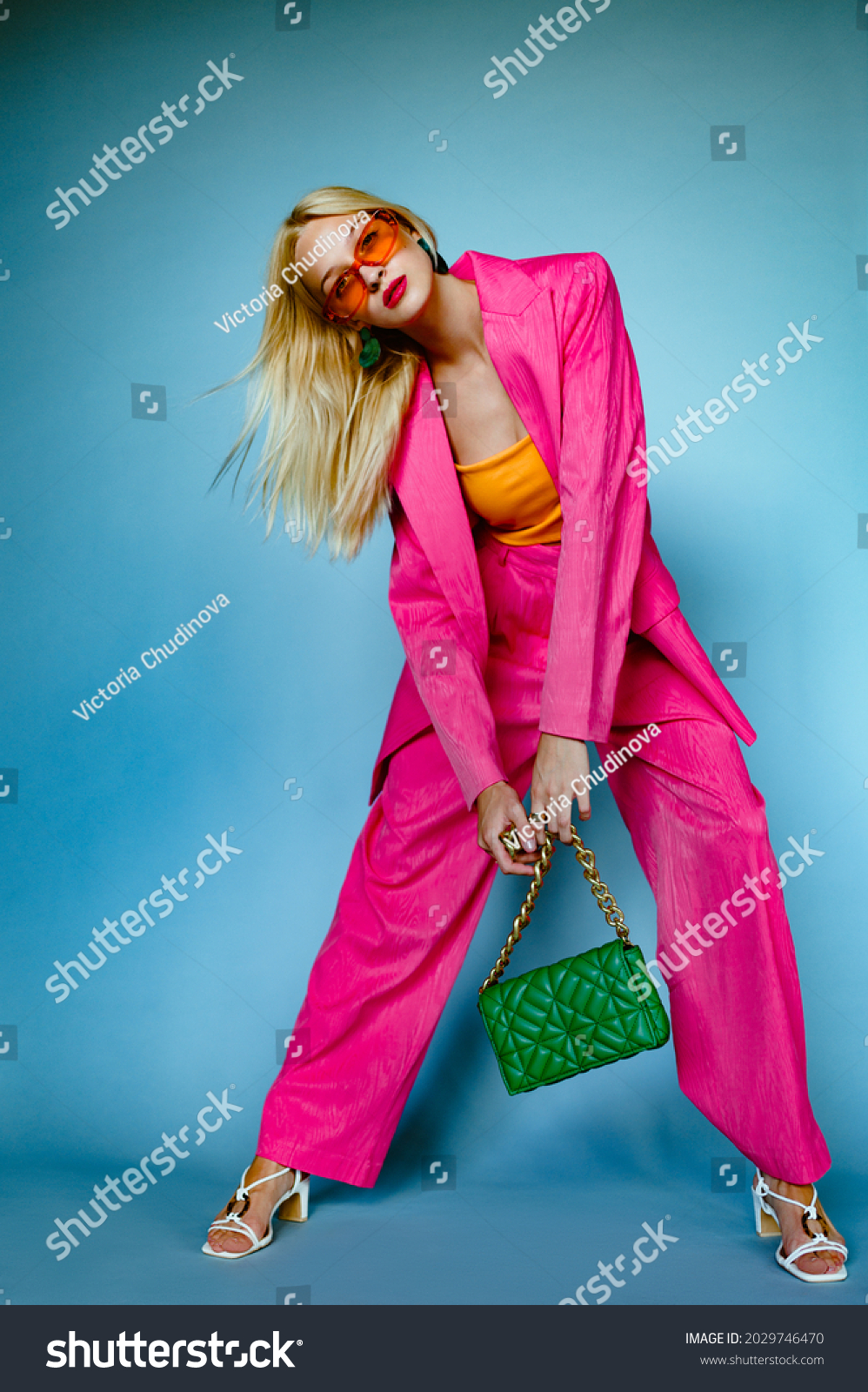 Fashionable woman wearing trendy fuchsia color suit. orange sunglasses, holding stylish green quilted faux leather bag with chunky chain, posing on blue background. Full-length studio fashion portrait #2029746470