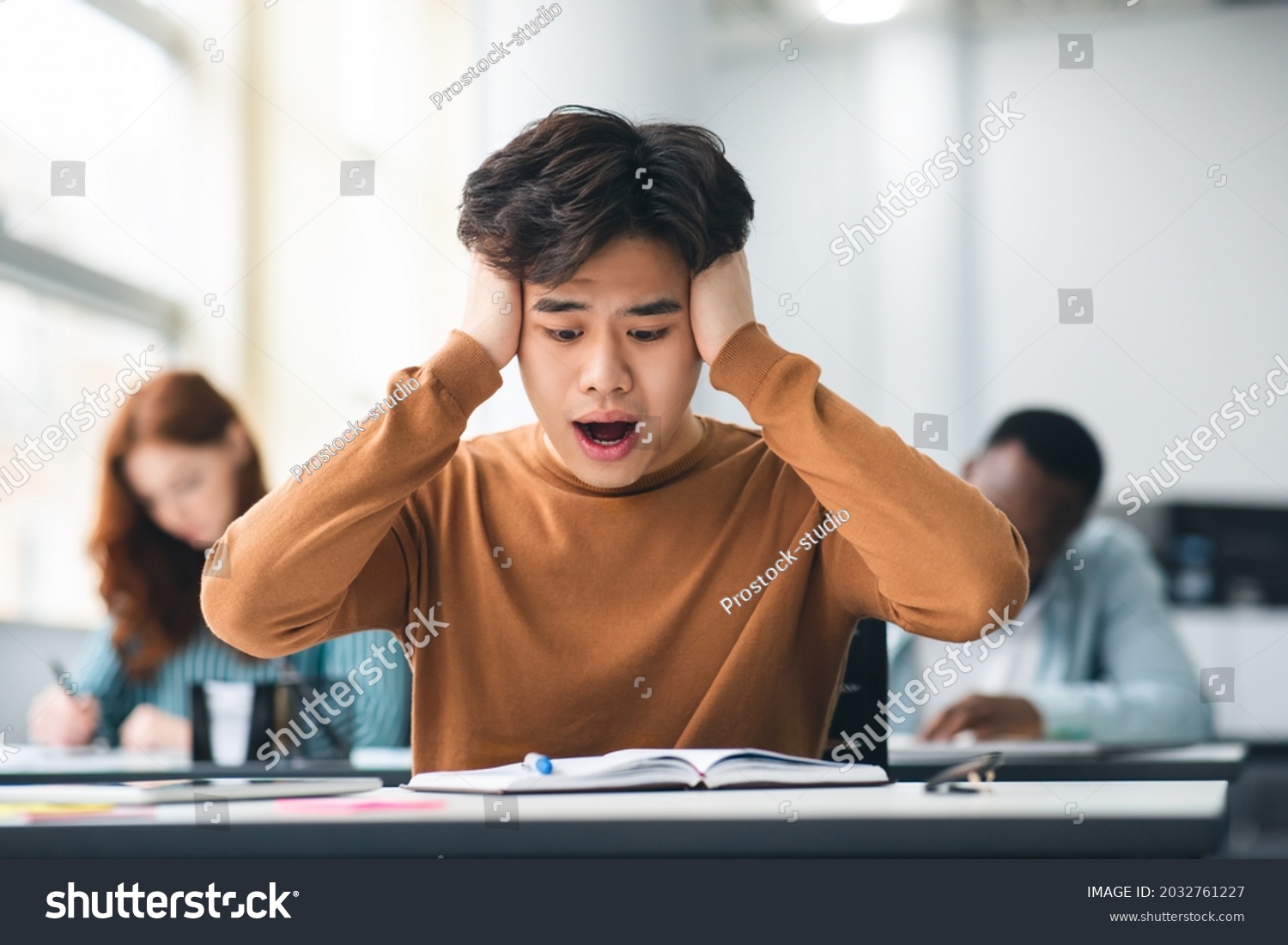 Portrait of shocked asian freshman sitting at table in classroom, grabbing clasping his head in horror and panic, looking at hard exam questions or test result, unhappy about deadline or low score #2032761227