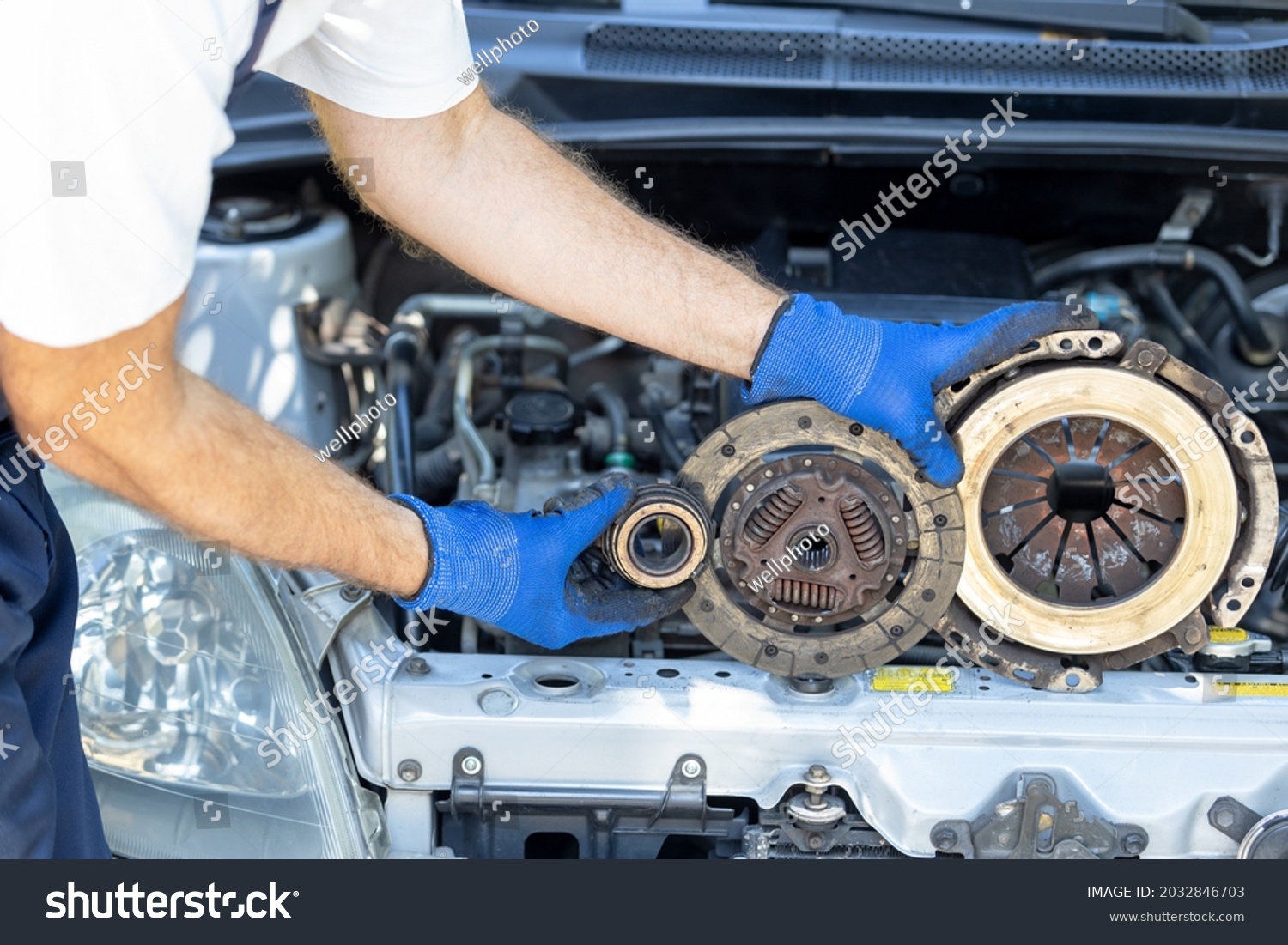 Automotive technician holding used car pressure plate, clutch disc and release bearing in front of the vehicle engine #2032846703