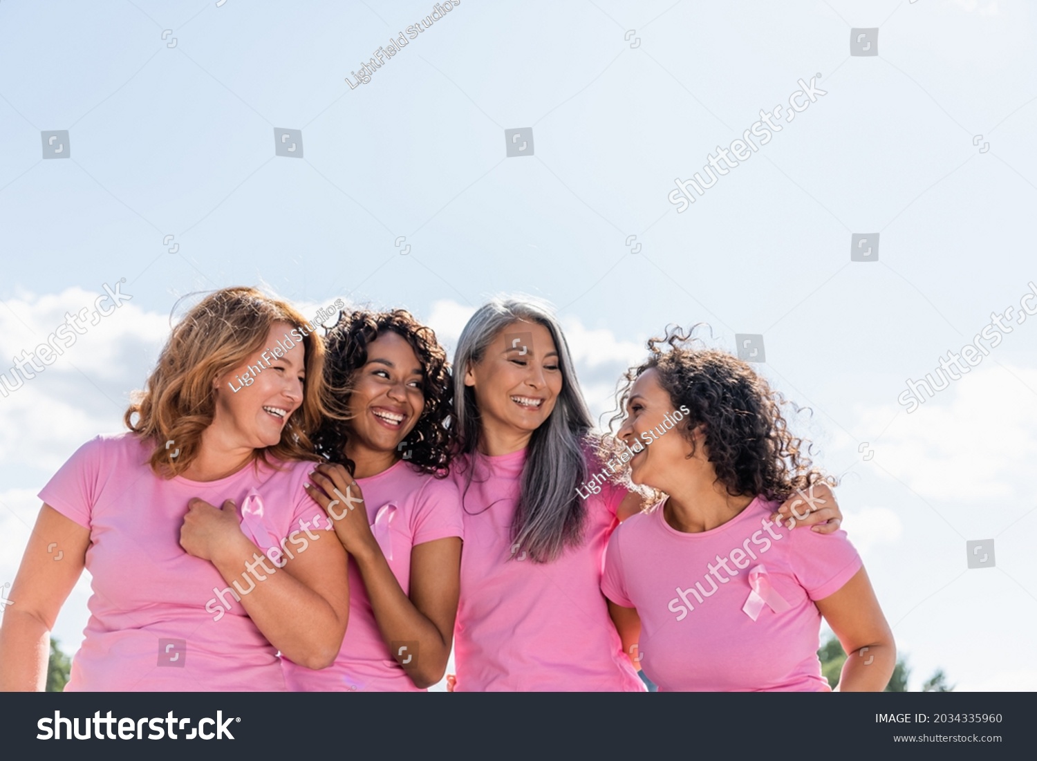 Cheerful multiethnic women with ribbons of breast cancer awareness hugging outdoors #2034335960