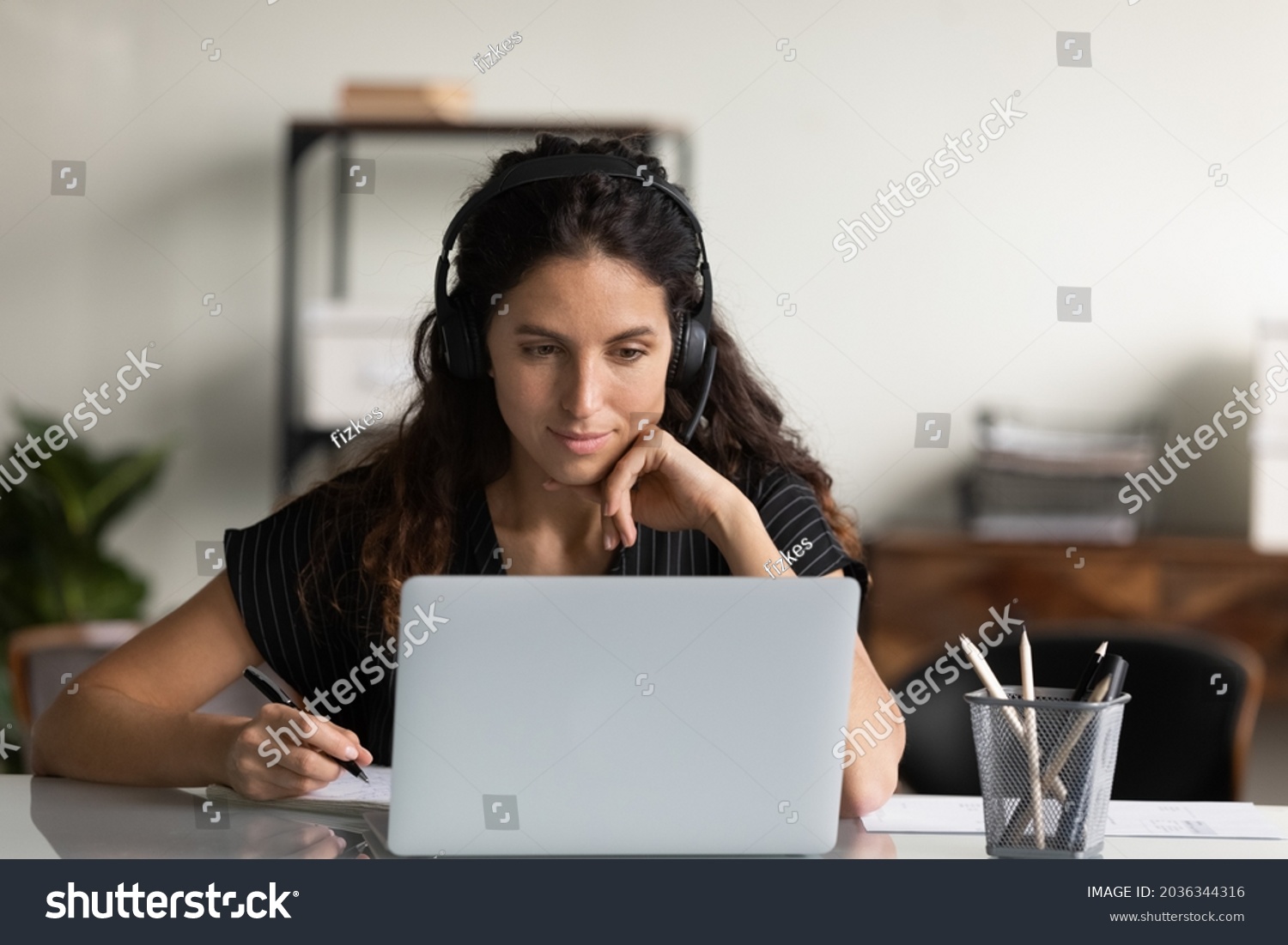 Focused female adult student in headphones using laptop, attending online lesson, virtual class, listening webinar, watching training, writing notes, making video call. Remote learning on internet #2036344316