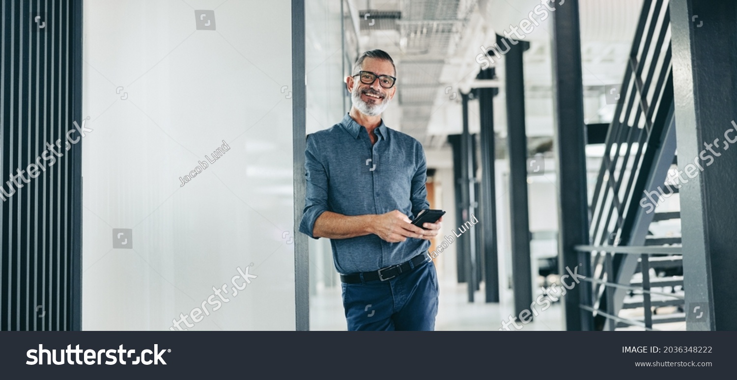 Smiling mature businessman holding a smartphone in an office. Businessman looking at the camera while standing alone in a modern workplace. Experienced businessman communicating with his clients. #2036348222