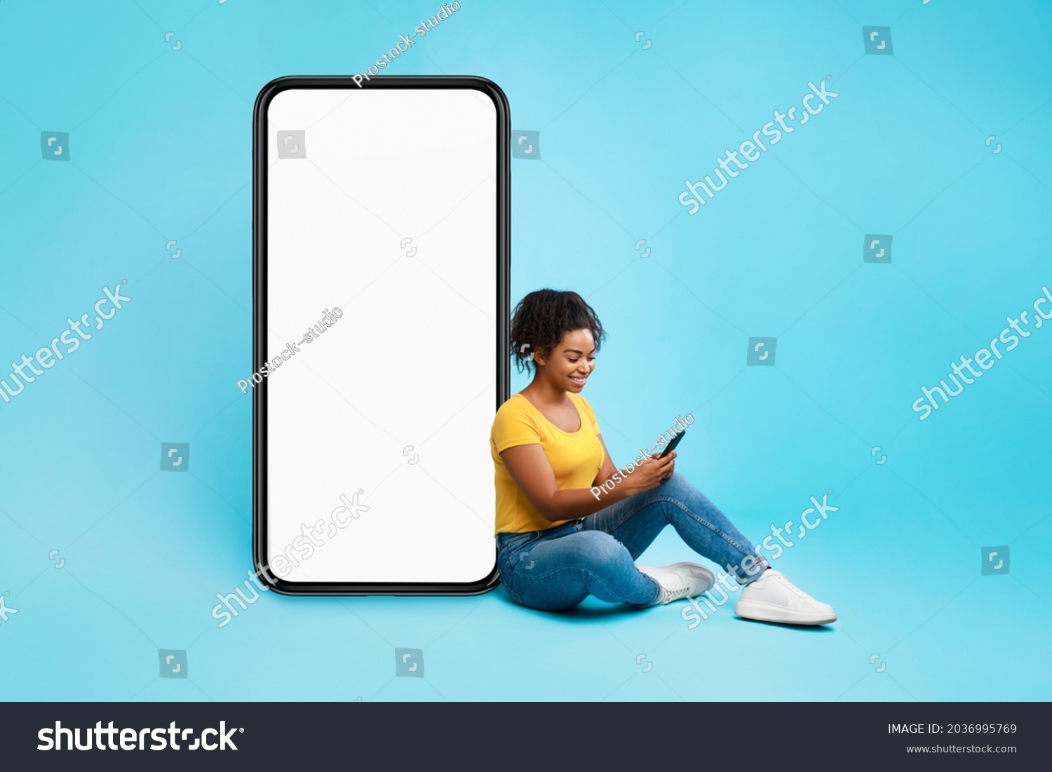 Cheery African American lady sitting near big cellphone with empty white screen, using mobile device, checking new cool app on blue studio background, mockup for website or application design #2036995769