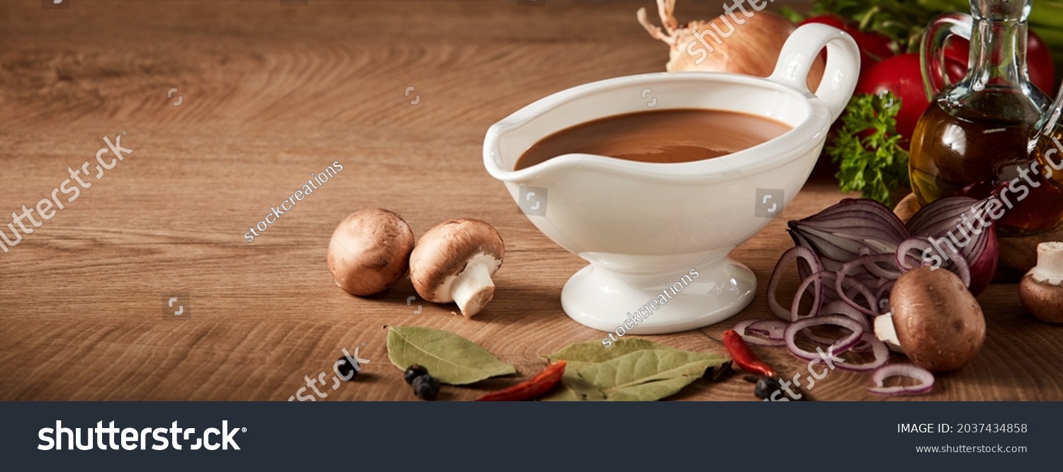 Sauce boat or sauciere filled with a rich brown gravy surrounded by fresh vegetable ingredients on a wood background with copyspace in panorama banner format #2037434858