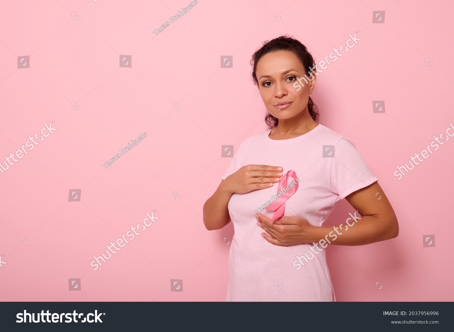 Mixed race woman puts hands around pink ribbon on her pink T Shirt, for breast cancer campaign, supporting Breast Cancer Awareness. Concept of 1 st October Pink Month and women's health care #2037956996