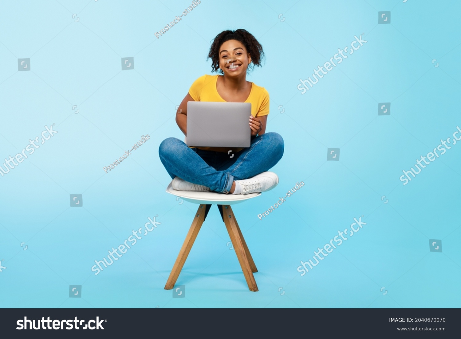 Cheery young black woman working online, sitting on chair and using laptop on blue studio background, full length. Cheerful African American lady surfing internet on portable pc #2040670070