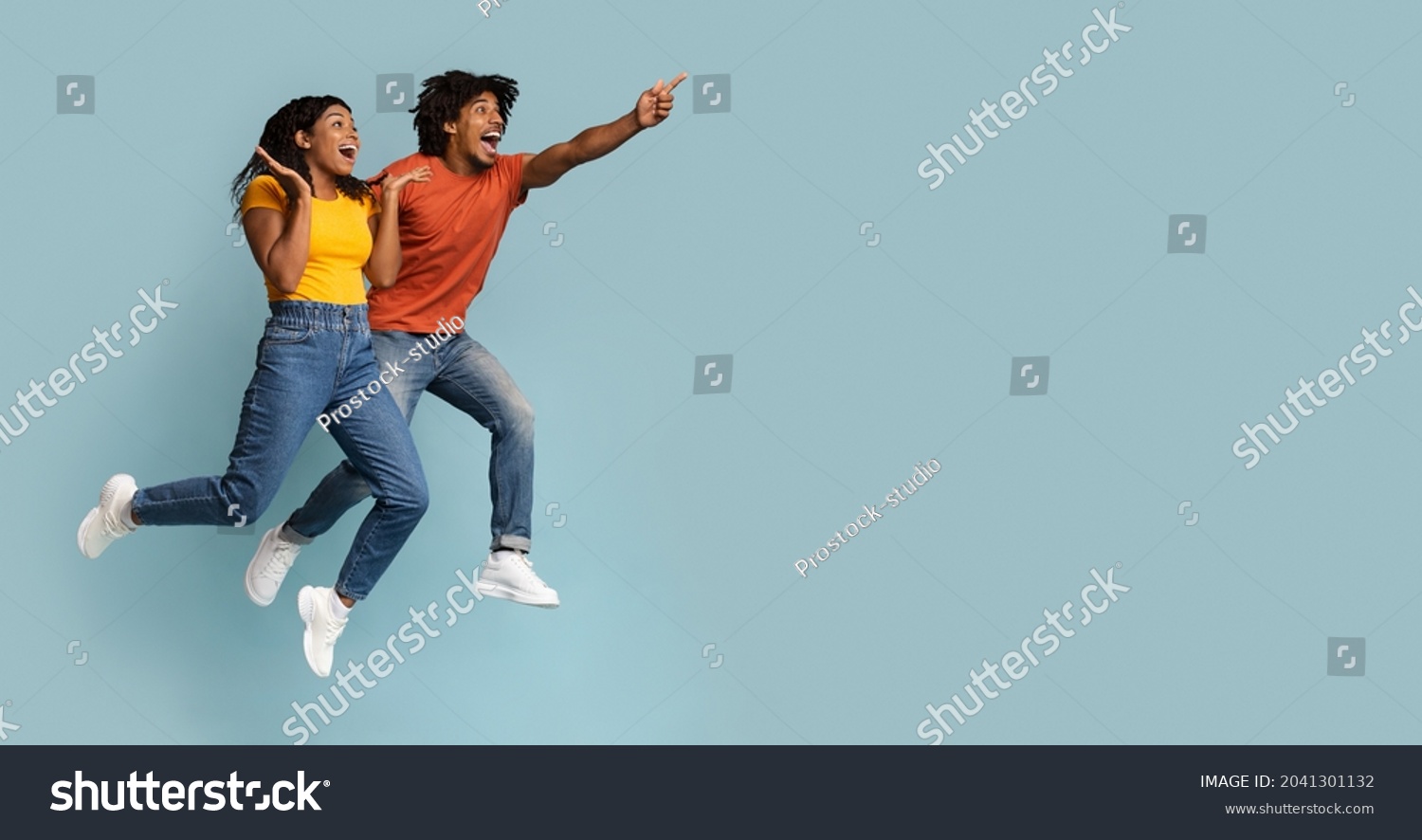 Carefree black millennial man and woman jumping up in the air and pointing at copy space, having fun on blue studio background, emotional couple running towards exciting advertisement, panorama #2041301132