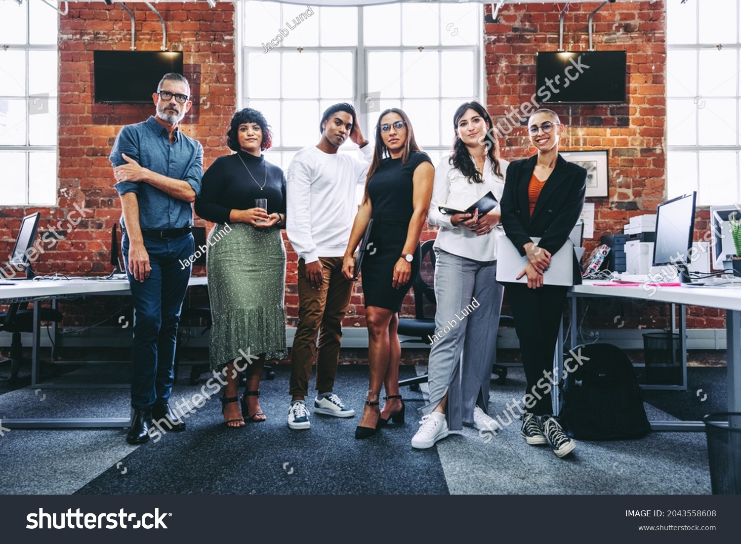 Diverse team of businesspeople standing together in an office. Group of modern businesspeople looking at the camera in a creative workplace. Business colleagues looking cheerful. #2043558608