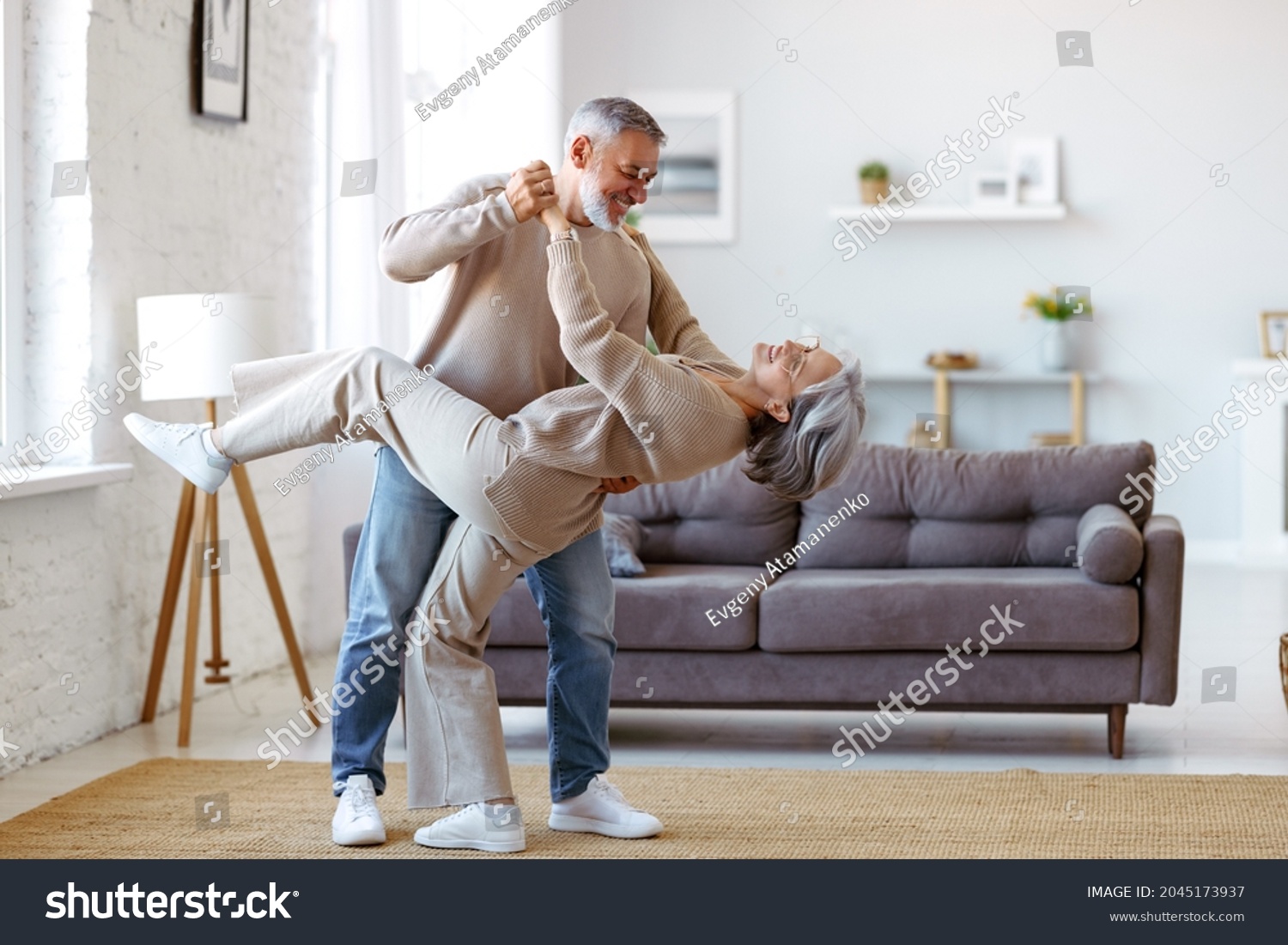 Keep moving. Romantic senior family couple wife and husband dancing to music together in living room, smiling laughing retired man and woman having fun, enjoying free time together at home #2045173937