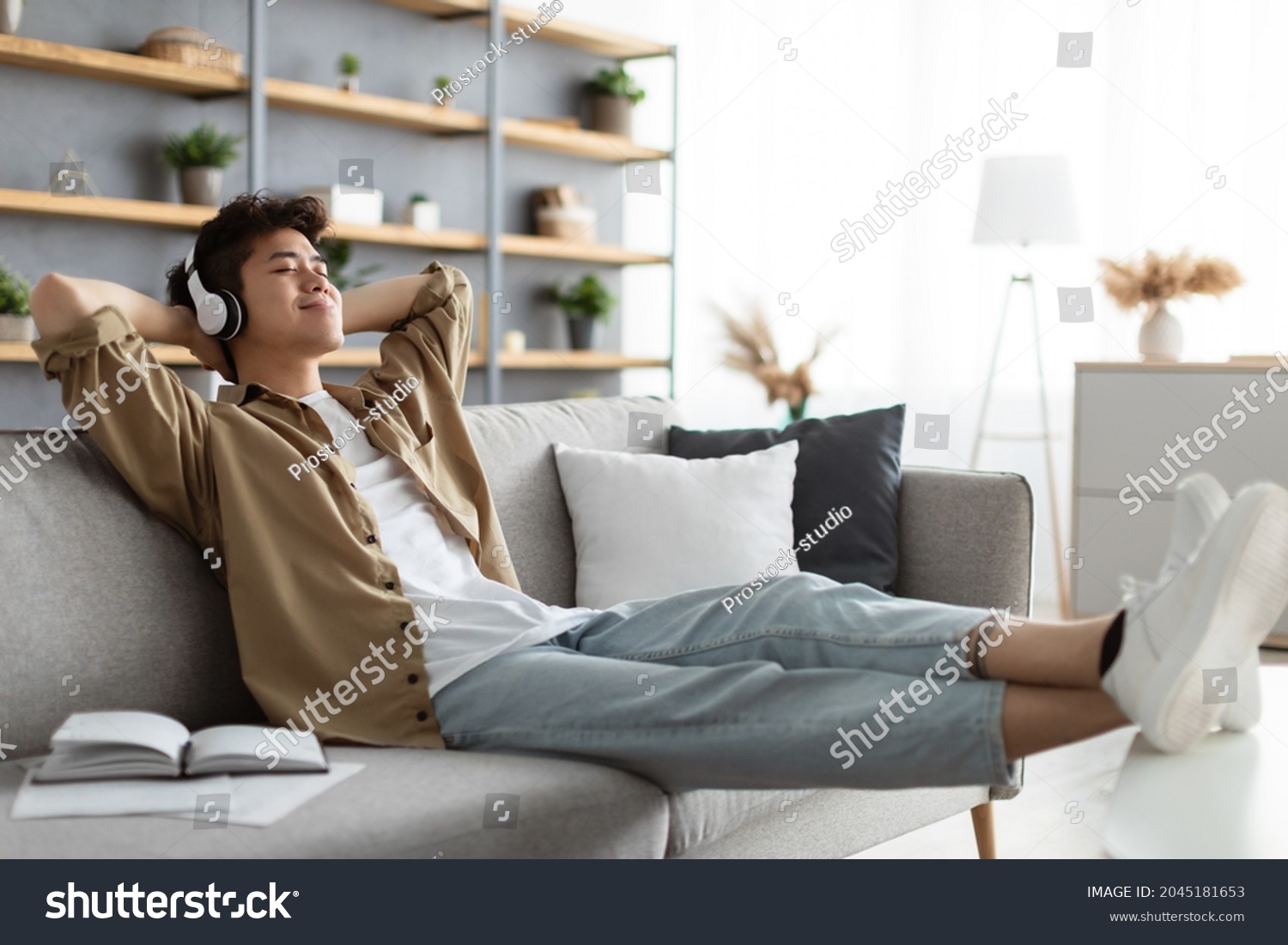 Rest And Relax Concept. Calm asian man sitting on couch, listening to music, audio book, podcast, enjoying meditation for sleep and peaceful mind in wireless headphones, leaning back, copy space #2045181653