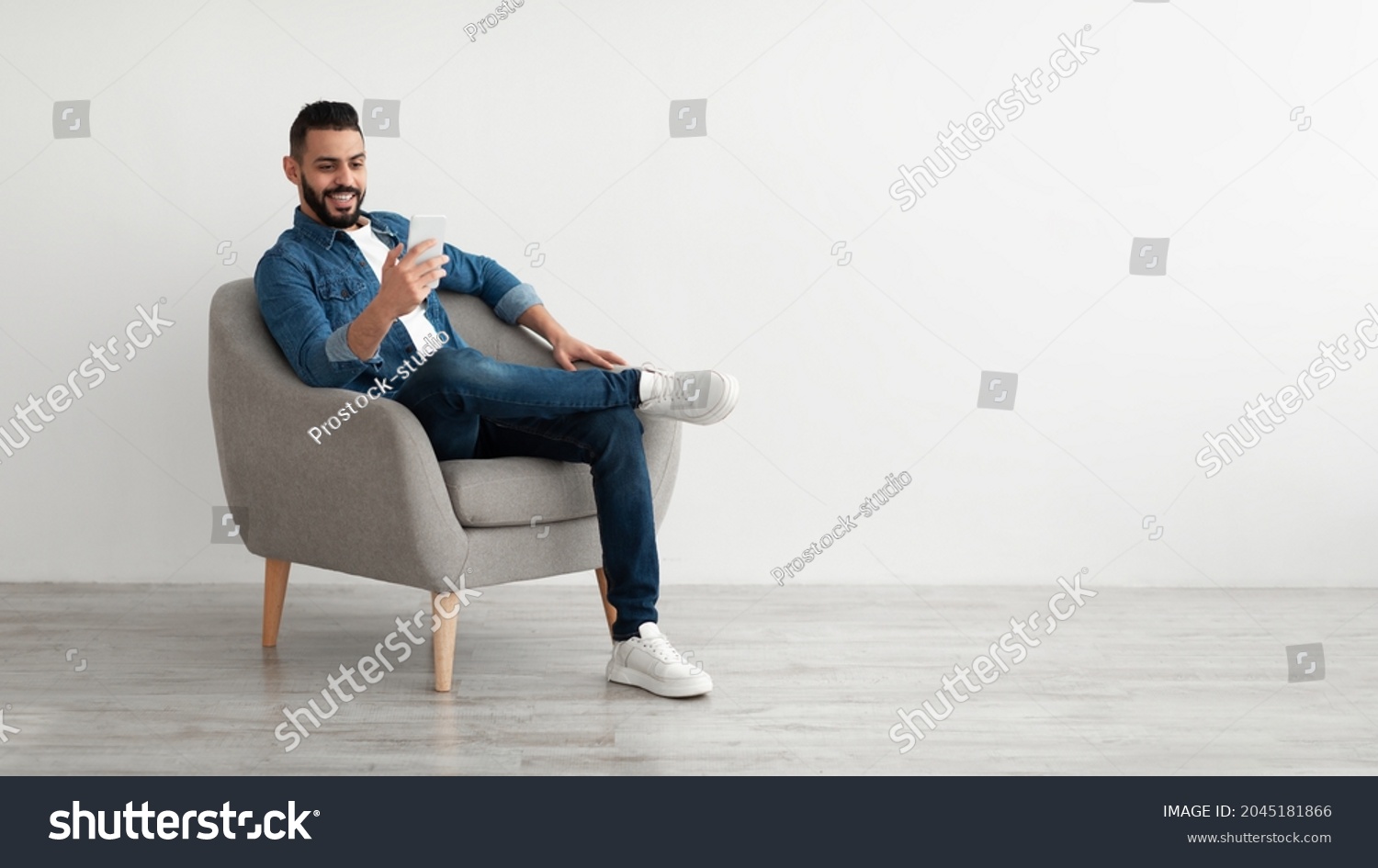 Cheerful young Arab man having online business meeting on smartphone, sitting in cozy armchair near white wall, banner design with free space. Eastern guy communicating remotely on mobile device #2045181866