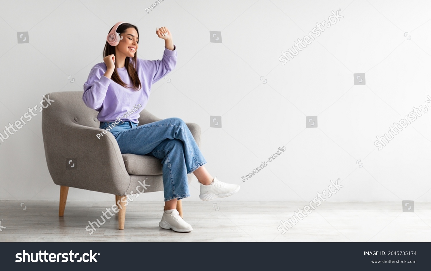 Cheery young lady in headphones listeing to music and dancing while sitting in armchair against white studio wall, banner design with free space. Carefree Caucasian woman moving to favorite song #2045735174